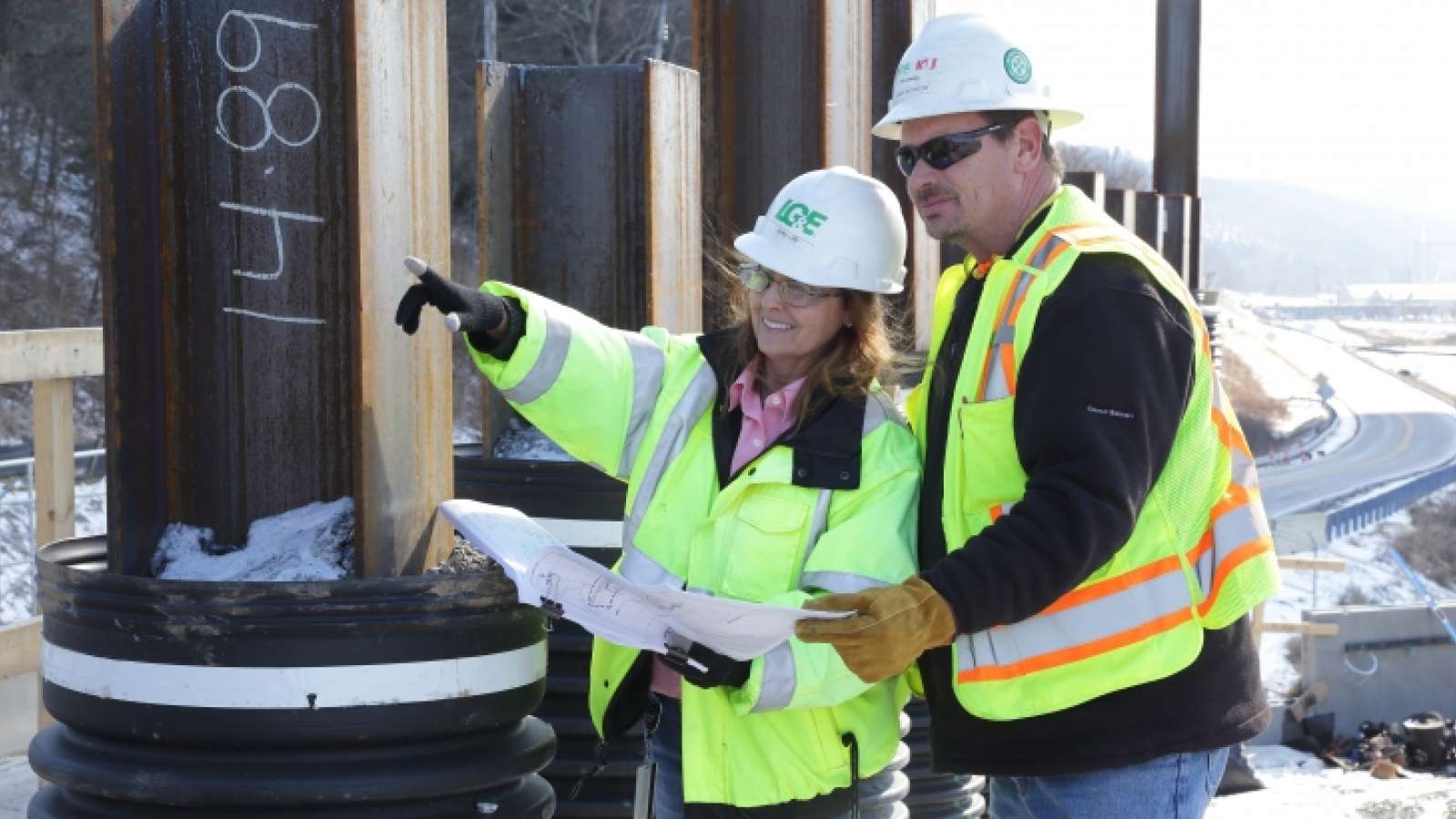 two employees with hard hats and safety vests looking at blueprints and pointing