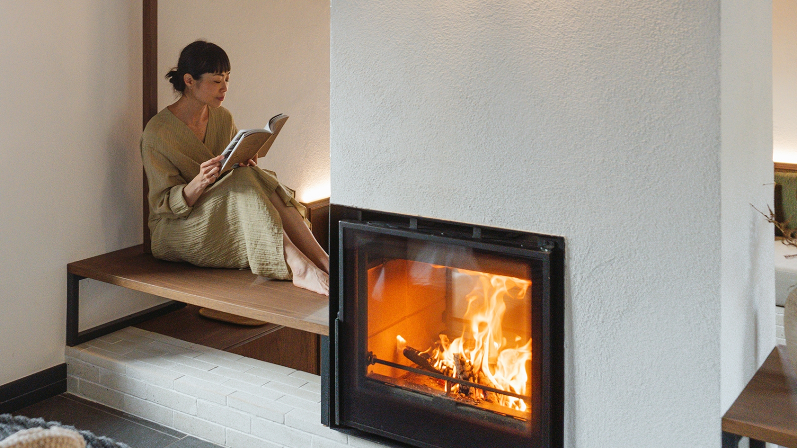 person at home reading a book on a bench next to a fireplace