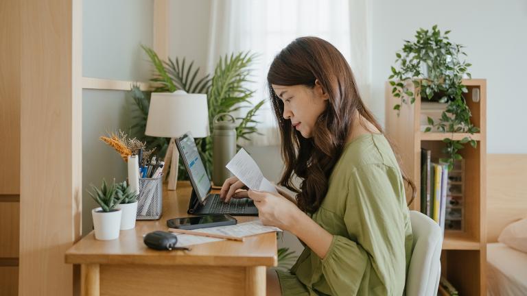 person sitting at a desk holding a bill and working on a laptop