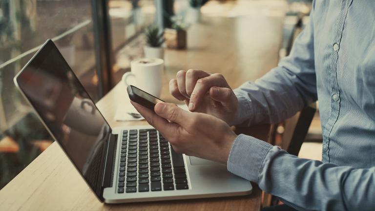 hands on a smartphone with a laptop in front on a table