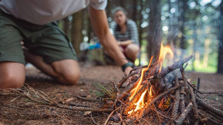 person in a forest starting a campfire while another person watches in the background