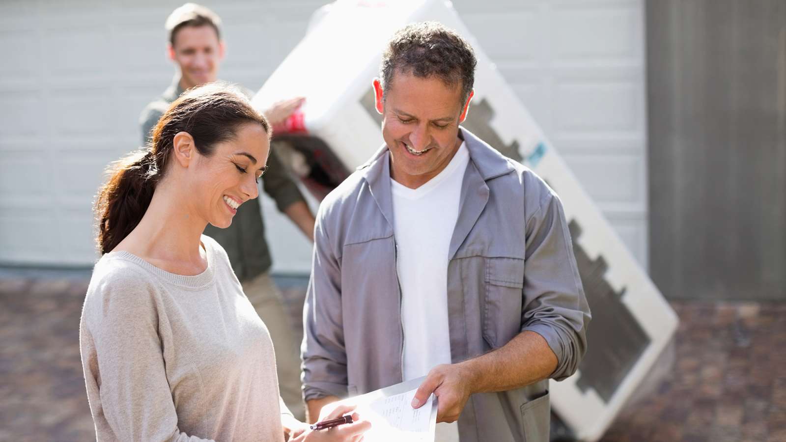 person signing a document while another person carries a refrigerator on a dolly
