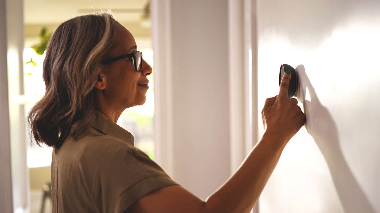 Side view of a person wearing glasses, adjusting a thermostat on an interior wall in a house.