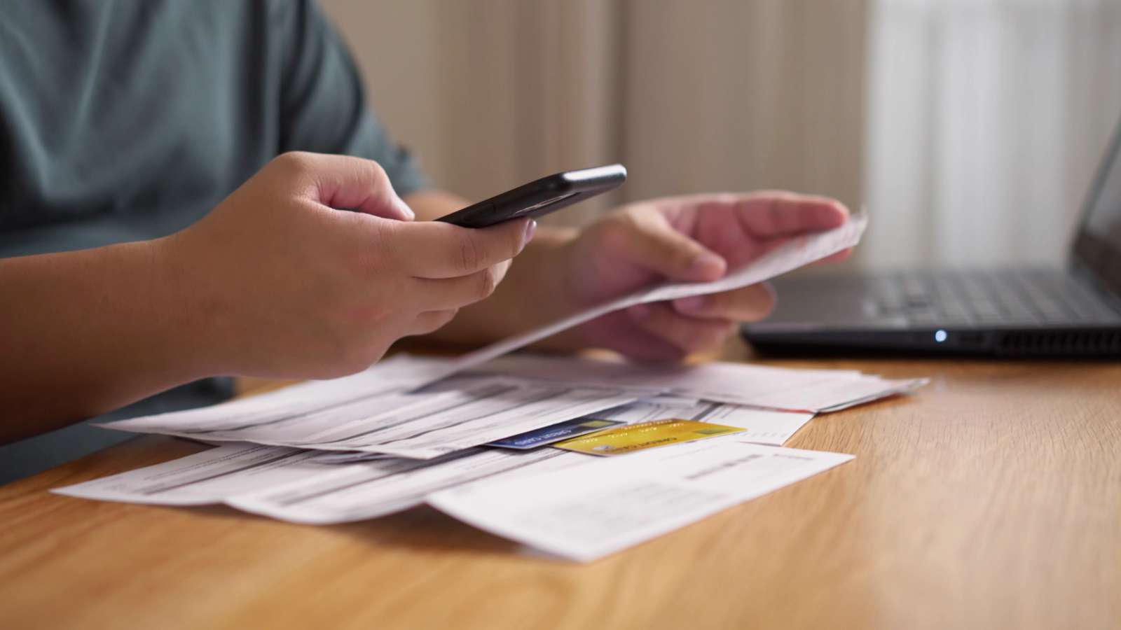 hands at a table with a laptop, a mobile phone, and paper bills