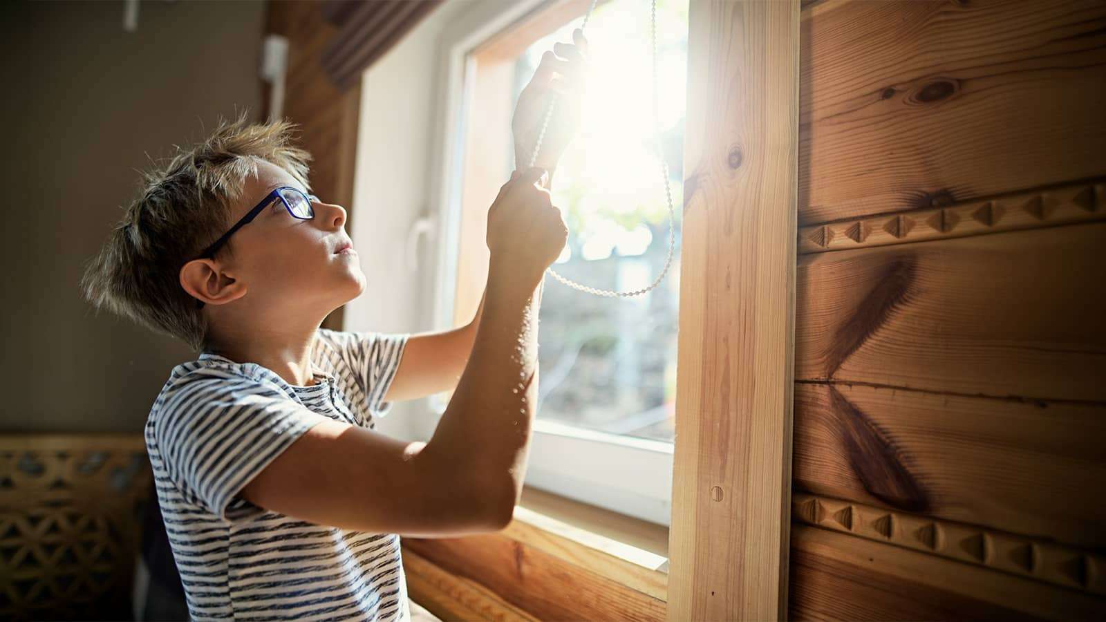 a boy wearing glasses lowering a window blind