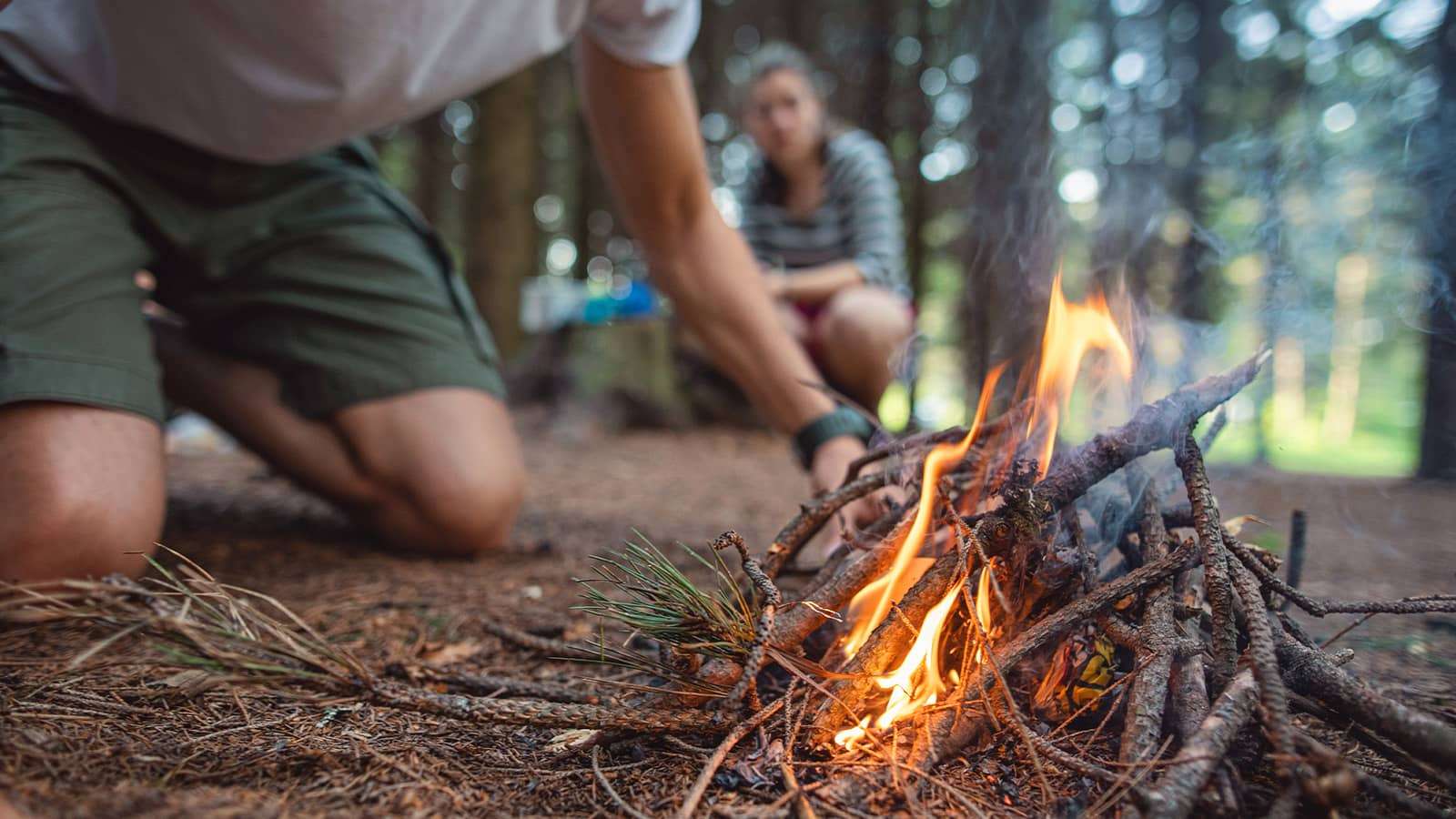 person in a forest starting a campfire while another person watches in the background